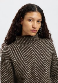 Pleteni pulover v rjavo-krem vzorcu ribje kosti, z visokim ovratnikom in teksturirano površino, z ohlapnim krojem in mehkim materialom.