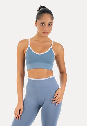 Kobieta stojąca, ubrana w niebieski bezszwowy sportowy biustonosz i legginsy z wysokim stanem z białym wykończeniem, neutralne tło.
