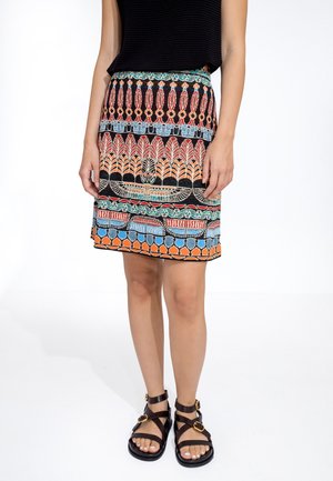 Multikleurige rok met ingewikkelde patronen in rood, blauw en oranje op een zwarte achtergrond; gecombineerd met bruine sandalen met bandjes.