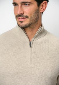 Beige stickad tröja med kvartdragkedja. Det strukturerade tyget visar ett subtilt mönster och en slät dragkedjeaccentee.