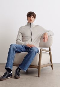 Grå zip-up jakke med høy krage, brukt sammen med blå jeans. Modellen sitter i en lys stoff-fotøyd lenestol med en tre-ramme.