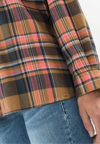 Camicia di flanella a quadri con colori marrone, arancione, rosa e blu navy. Presenta un'etichetta a linguetta vicino all'orlo e polsini conbottoni. Indossata con jeans blu.