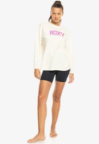 Mujer de pie descalza, vistiendo una camiseta de manga larga color crema con "ROXY" en rosa y negro, y pantalones cortos ajustados, contra un fondo blanco.