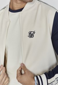 Šviesiai rudas bomberis su tamsiai mėlynu siuvinėtu logotipu ant krūtinės, užtrauktuku užsegamas, su gipiūro apykakle ir kontrastingomis tamsiai mėlynomis rankovėmis su juostelėmis.