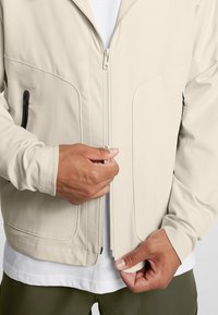 Krämfärgad, lätt jacka med dragkedja fram, med sidofickor med svarta detaljer och synliga sömmar.