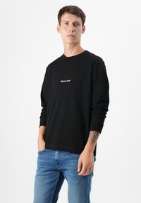Jonge man met kort haar, gekleed in een zwart longsleeve shirt en blauwe jeans, staat tegen een effen witte achtergrond en kijkt iets buiten de camera.