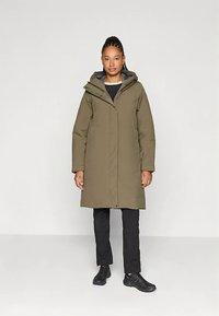 Olivovo zelený dlhý kabát s kapucňou, vybavený plným zipsom a zapínaním na cvočky, nosený cez čierne nohavice a topánky. Hladká textúra, minimalistický dizajn.