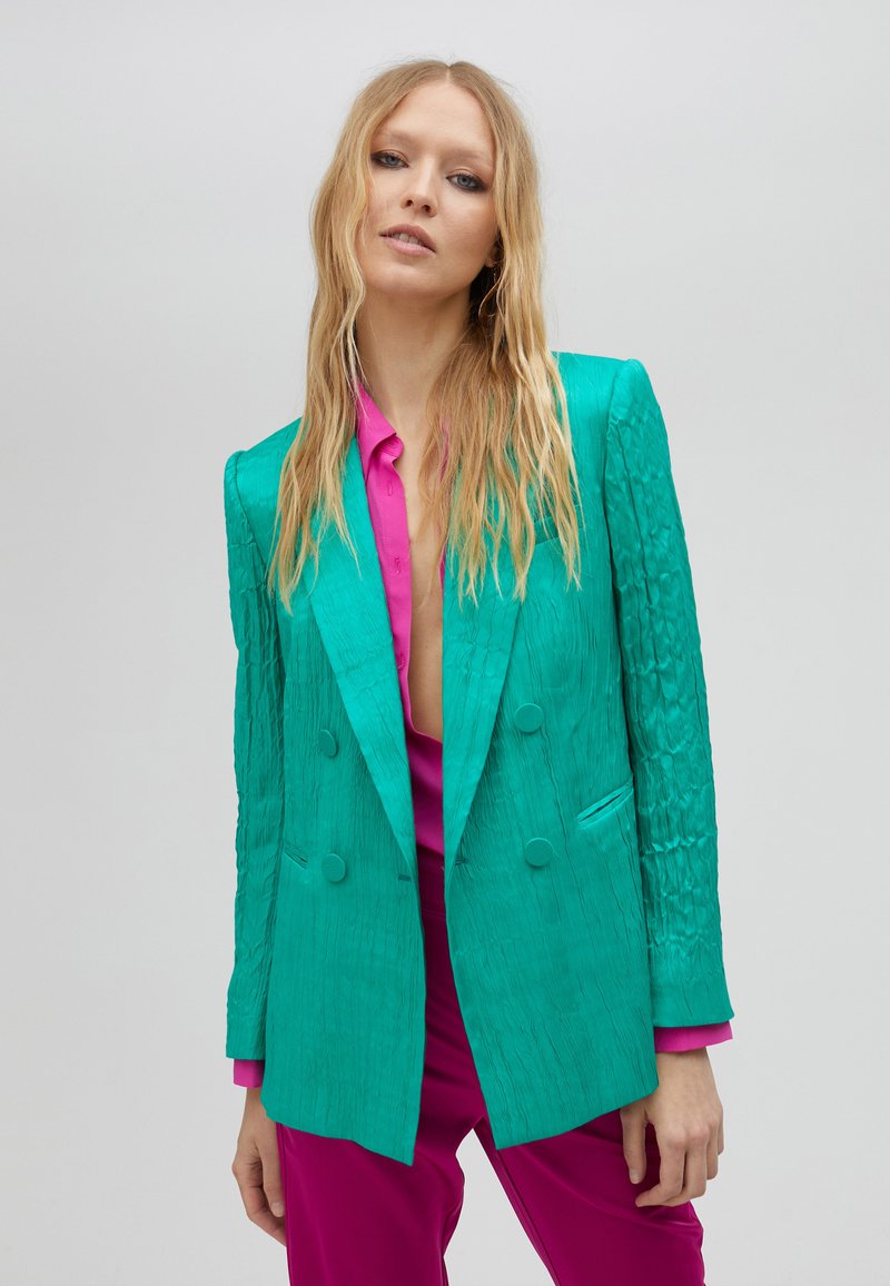 Blazer de doble botonadura en turquesa con tejido texturizado, solapas con muesca y dos bolsillos frontales, combinado con una camisa rosa vibrante.