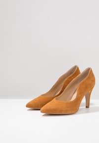 Semišové lodičky v hořčicově žluté barvě s špičatou špičkou a V-řezem v přední části; mají tenký podpatek pro zvýšení výšky.