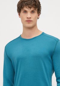 Camisa de manga longa turquoise com decote redondo, apresentando uma textura fina e costuras em contraste ao longo das bordas.