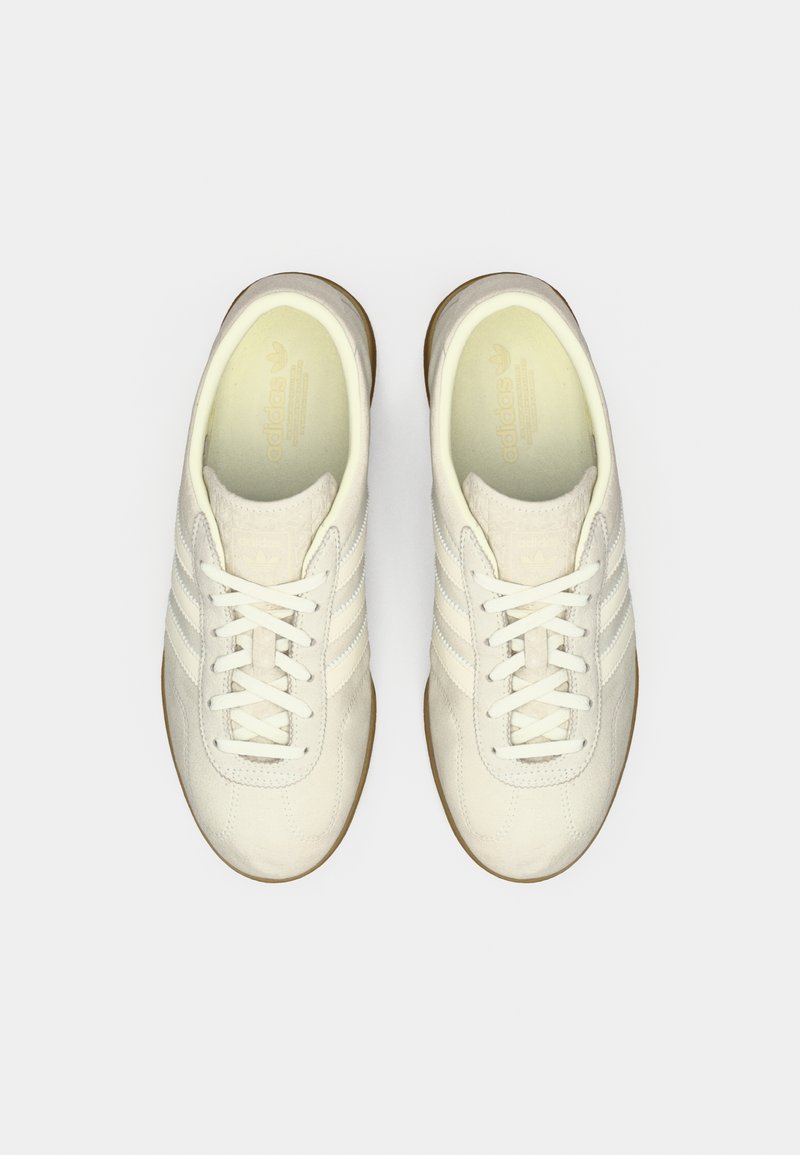 White leather sneakers with three side stripes, round toe, lace-up design, and gum rubber outsole. Smooth texture and minimalist style.