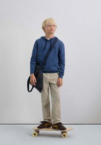 Sudadera con capucha azul con cordón, pantalones beige y zapatillas marrones sobre una patineta. Bolsa mensajera negra colgada del hombro. Textura de madera suave.