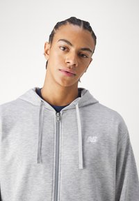 Jaunas žmogus su pintomis plaukų kasytėmis, vilkintis šviesiai pilką užtrauktuku megztinį su "New Balance" logotipu, šiek tiek pasviręs į priekį prieš paprastą foną.