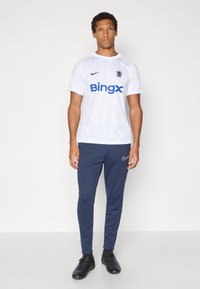 Hvit sportsdrakt med blå "Bingx" logo, korte ermer og subtilt teksturmønster. Kombinert med marineblå treningsbukser og svarte sko.