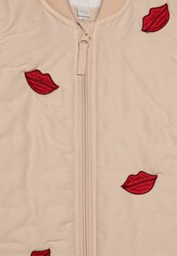 Lys beige bomberjakke med lynlås foran, der har røde broderede læber som accenter og et struktureret, let pufet stof.