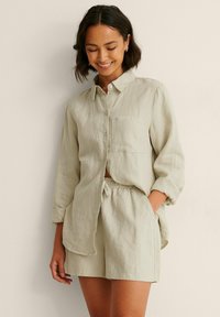 Light beige linen shirt with long sleeves and a chest pocket, paired with matching shorts featuring an elastic waistband. Soft texture.
