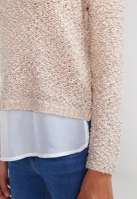 Pull en maille beige clair avec une finition texturée, présentant des manches longues et un design superposé sur un haut blanc transparent.