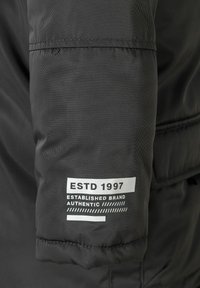 Morbida giacca nera con maniche imbottite e testo bianco "ESTD 1997" e "Marchio Autentico Fondato" stampato vicino al polsino.