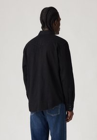 Camisa preta de manga comprida com punhos abotoados e design de pala, combinada com jeans de ganga azul. Apresenta um corte relaxado e textura suave.