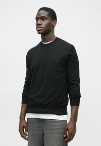 Jovem vestido com uma camisa preta de mangas longas sobre uma camiseta branca e jeans cinzentos, de pé contra um fundo branco liso.