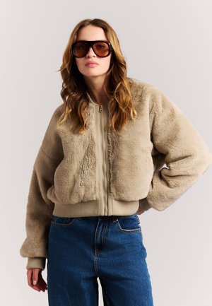 Vrouw met golvend haar draagt grote getinte zonnebril, een beige pluizige jas met rits en blauwe spijkerbroek, staand met één hand op haar heup.