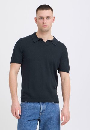 Jeune homme aux cheveux courts portant un polo noir à manches courtes et un jean bleu, debout devant un fond clair uni.