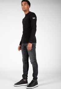 Maglietta nera a maniche lunghe con logo sulla manica sinistra, abbinata a jeans slim-fit grigio scuro e sneakers nere con suola bianca.