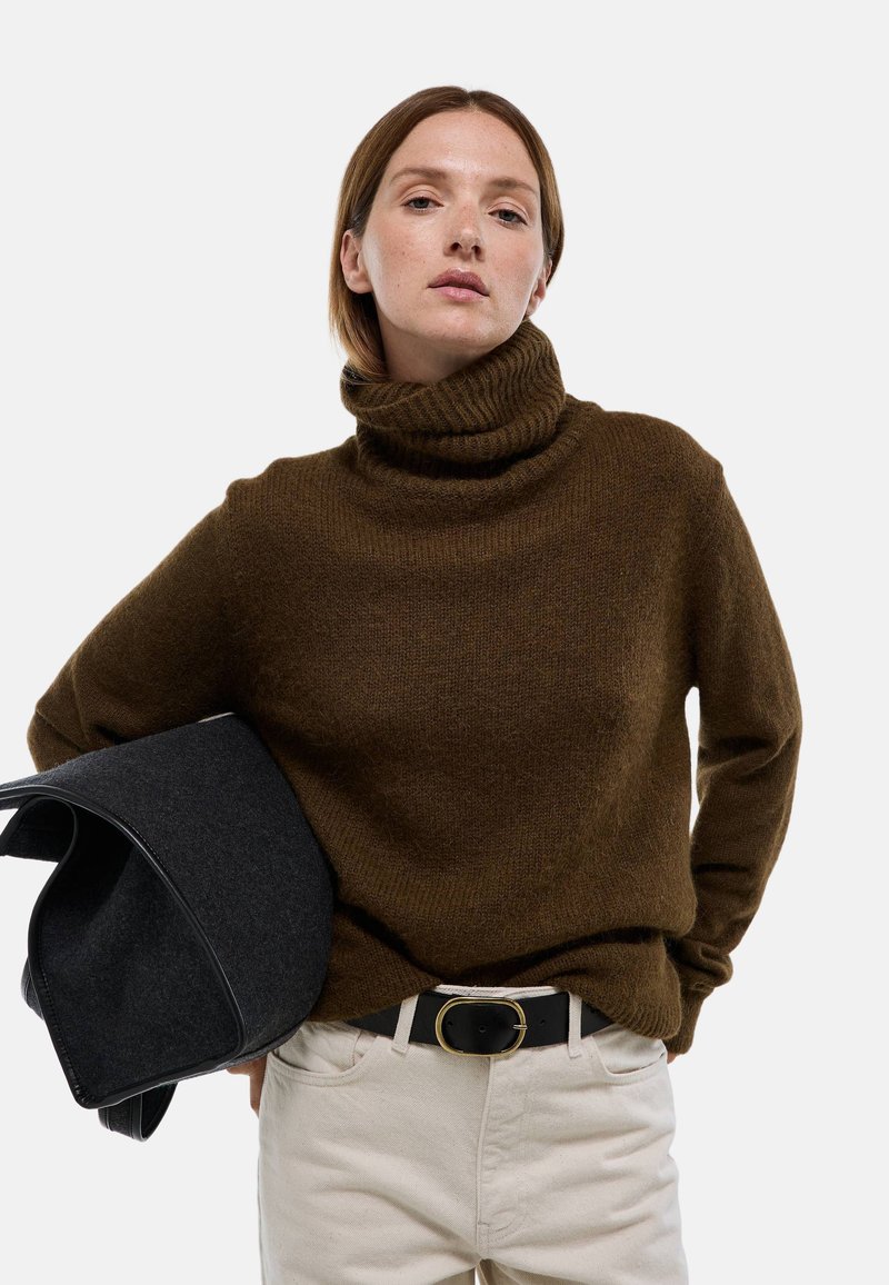 Pull à col roulé marron en maille douce, présentant un col côtelé et une coupe décontractée, associé à un pantalon crème et un sac noir.