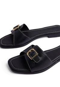 Sandalias deslizables de cuero negro con punta cuadrada, que presentan un acento de hebilla dorada y costuras en blanco que contrastan a lo largo de los bordes. Suela plana.