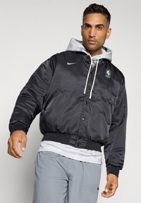 Mustat satiinibomberitakki hupulla, Nike-logo, NBA-patchi, edessä napit, ribbatut hihansuut ja sivutaskut. Päällä valkoinen paita.