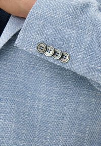 Tessuto azzurro chiaro a zigzag con una superficie liscia. Quattro bottoni circolari grigi con due fori ciascuno sul polsino.