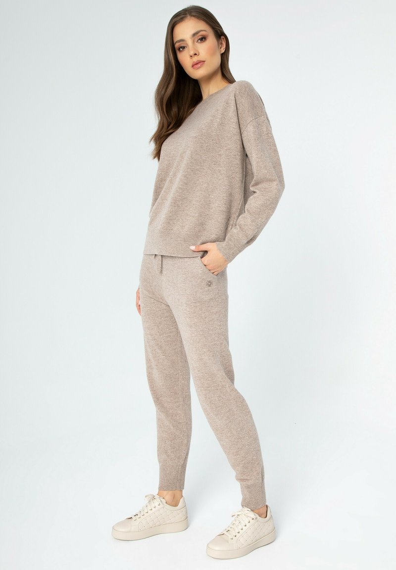 Beige Loungewear-Set aus weichem Strickmaterial, bestehend aus einem lässigen Sweater und tapered Hosen mit elastischem Bund. Mit dezentem Branding akzentuiert.
