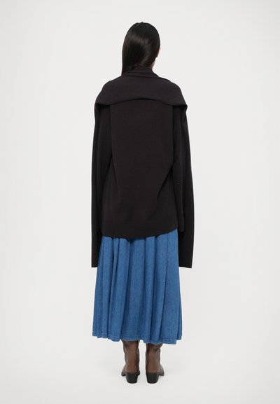 Pull en maille noir surdimensionné avec des manches larges, associé à une longue jupe en denim bleue. L'ensemble est complété par des bottes à talons marron.