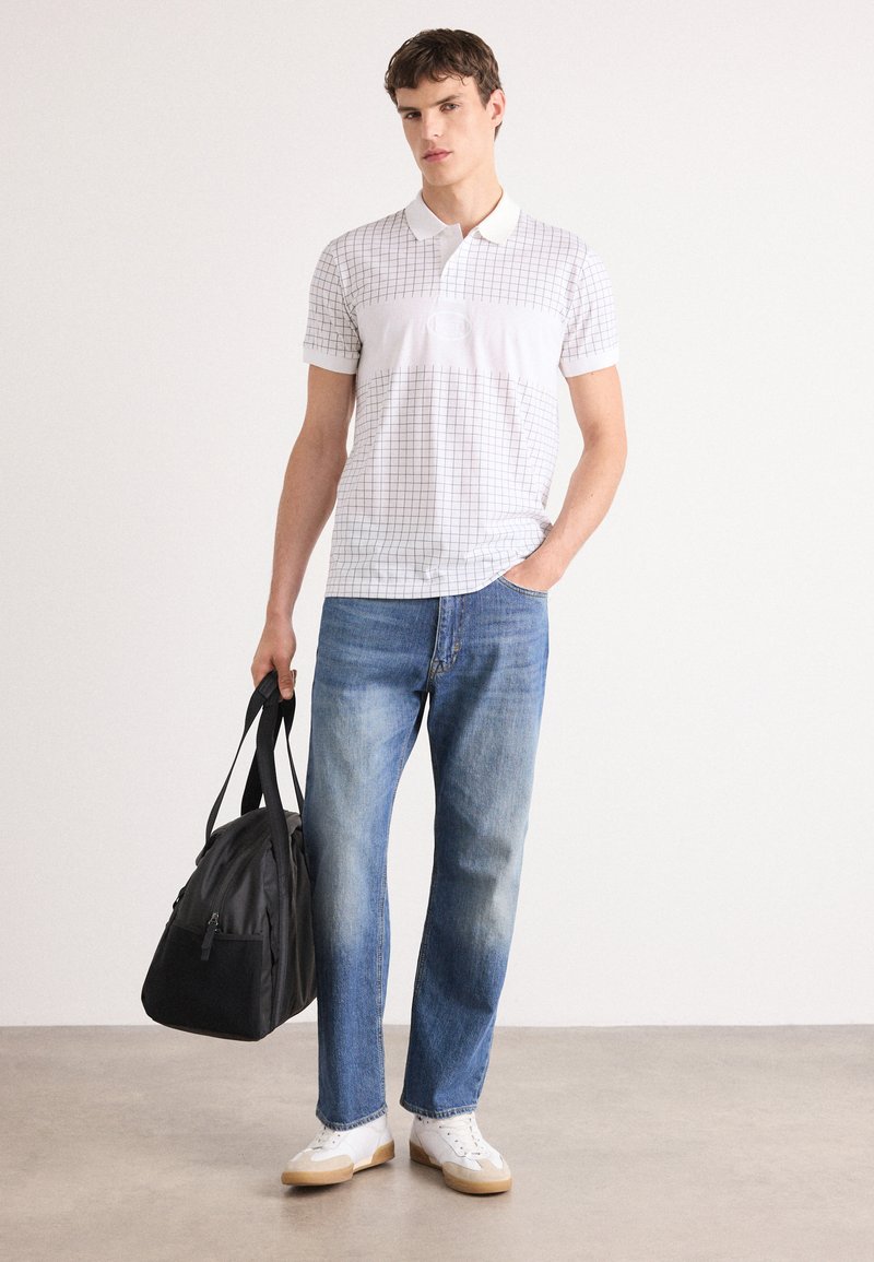 Camisa polo blanca con un patrón de cuadros, combinada con jeans de color azul claro y zapatillas blancas. Sostenido en una mano hay un bolso de deporte negro.