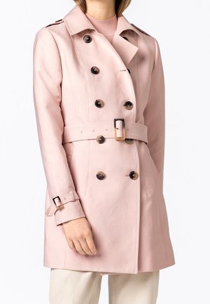 Mujer con un abrigo trench doble botonadura en rosa claro, con cinturón y detalles de hebilla, combinado con pantalones beige.