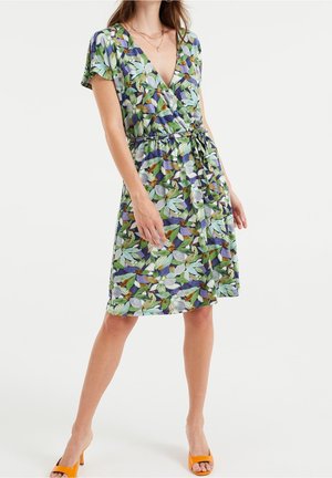 Woman wearing a knee-length, short-sleeve floral wrap dress with blue, green, and white leaves and orange heeled sandals.