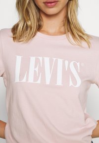Ljusrosa bomulls-T-shirt med rundad halsringning, med den djärva vita "LEVI'S"-logotypen över bröstet. Mjukt material med korta ärmar.