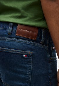 Κοντινό πλάνο ατόμου που φοράει σκούρο μπλε τζιν Tommy Hilfiger με καφέ δερμάτινο λογότυπο και πράσινο πουκάμισο μέσα στο παντελόνι.