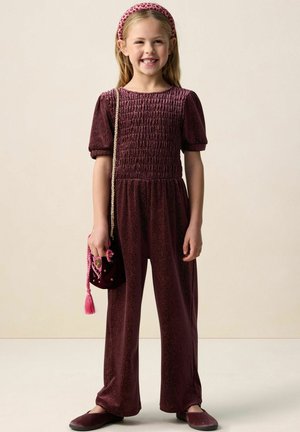 Fille souriante portant une combinaison texturée bordeaux, un bandeau rose, tenant un petit sac assorti avec un pompon, debout sur un fond uni.