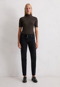 Vrouw leunend tegen een witte muur, gekleed in een strakke, bruine, korte mouwen turtleneck, zwarte drawstring broek en zwarte instappers.