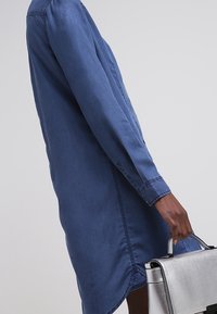 Abito in denim blu con colletto, maniche lunghe e spacchi laterali; abbinato a una borsa argentata lucida.
