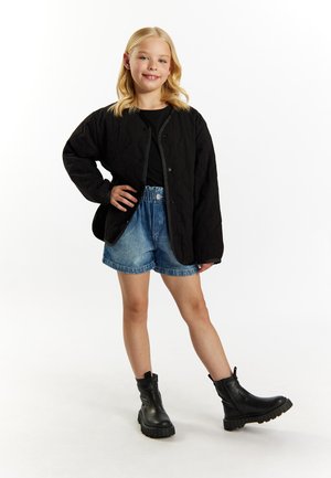 Mustat vuoratut takki, jossa on pyöreä helma, farkkushortsit ja mustat nilkkasaappaat paksuilla pohjilla. Malli poseeraa yksi käsi lantiolla.