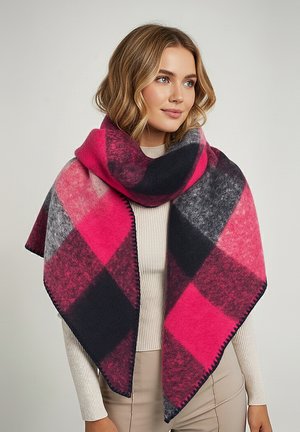 Woman wearing a large pink, black, and gray checkered wool scarf over a cream ribbed sweater and beige pants against a plain background.