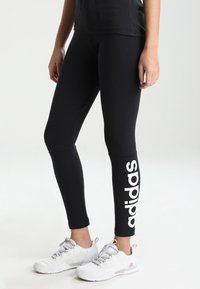 Černé legíny s výrazným bílým logem "adidas" podél levé nohy, doplněné bílými sportovními botami a černým topem.