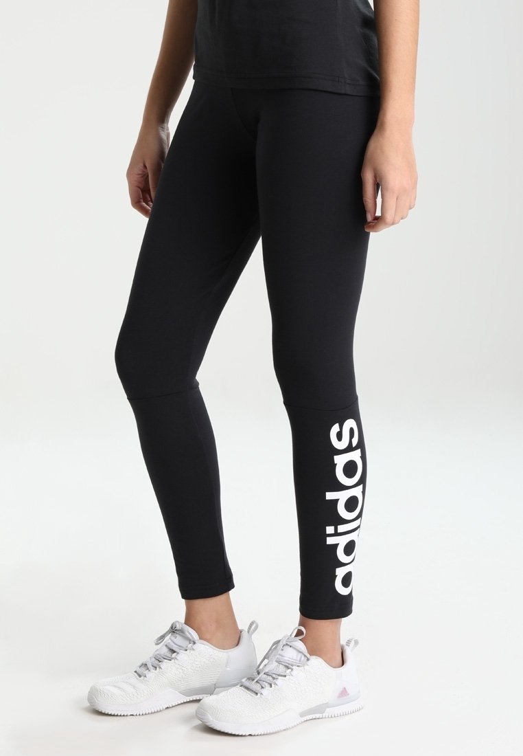 Černé legíny s výrazným bílým logem "adidas" podél levé nohy, doplněné bílými sportovními botami a černým topem.