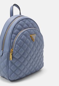 Kleine gewatteerde lichtblauwe rugzak met gouden ritsen en driehoekig Guess-logo op het voorvak, afgebeeld tegen een effen lichte achtergrond.