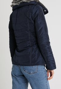 Giubbotto puffer blu navy con motivo trapuntato, dotato di cappuccio foderato in pelliccia sintetica e polsini elasticizzati. Indossato sopra a jeans blu.