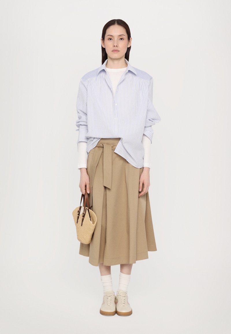 Chemise oversized rayée bleu et blanche, jupe portefeuille beige, baskets crèmes, chaussettes blanches et petit sac tissé.