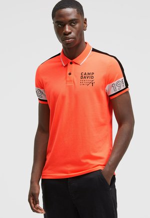 Bright orange polo shirt with black and white accents, featuring a collar, short sleeves, and printed branding on the chest and sleeve.