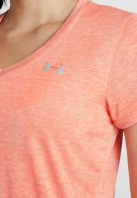 Camiseta de manga corta con escote en V en color coral brillante, confeccionada en un tejido texturizado con un sutil patrón jaspeado. Presenta un pequeño logo gris.